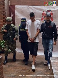 En la foto aparece en un pasillo, en acción de desplazamiento, con los brazos adelante y esposado, Víctor Manuel Rivas España. Viste Camiseta blanca, de manga corta; pantalón corto oscuro y zapatillas blancas. Al lado derecho de él hay un uniformado de la Policía y del Ejército. Al lado izquierdo hay un servidor del CTI de la Fiscalía. Detrás de ello se ve un pendón institucional del Ejército.