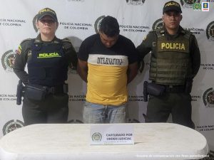 En la fotografía aparece un hombre capturado, acompañado de dos uniformados de la Policía. En la parte posterior un banner con logos de la entidad.   