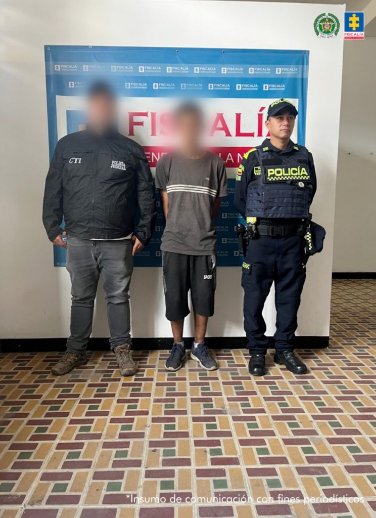El imputado se encuentra custodiado por un servidor del Cuerpo Técnico de Investigación CTI y un uniformado de la Policía Nacional. Detrás se observa un backing de la Policía.