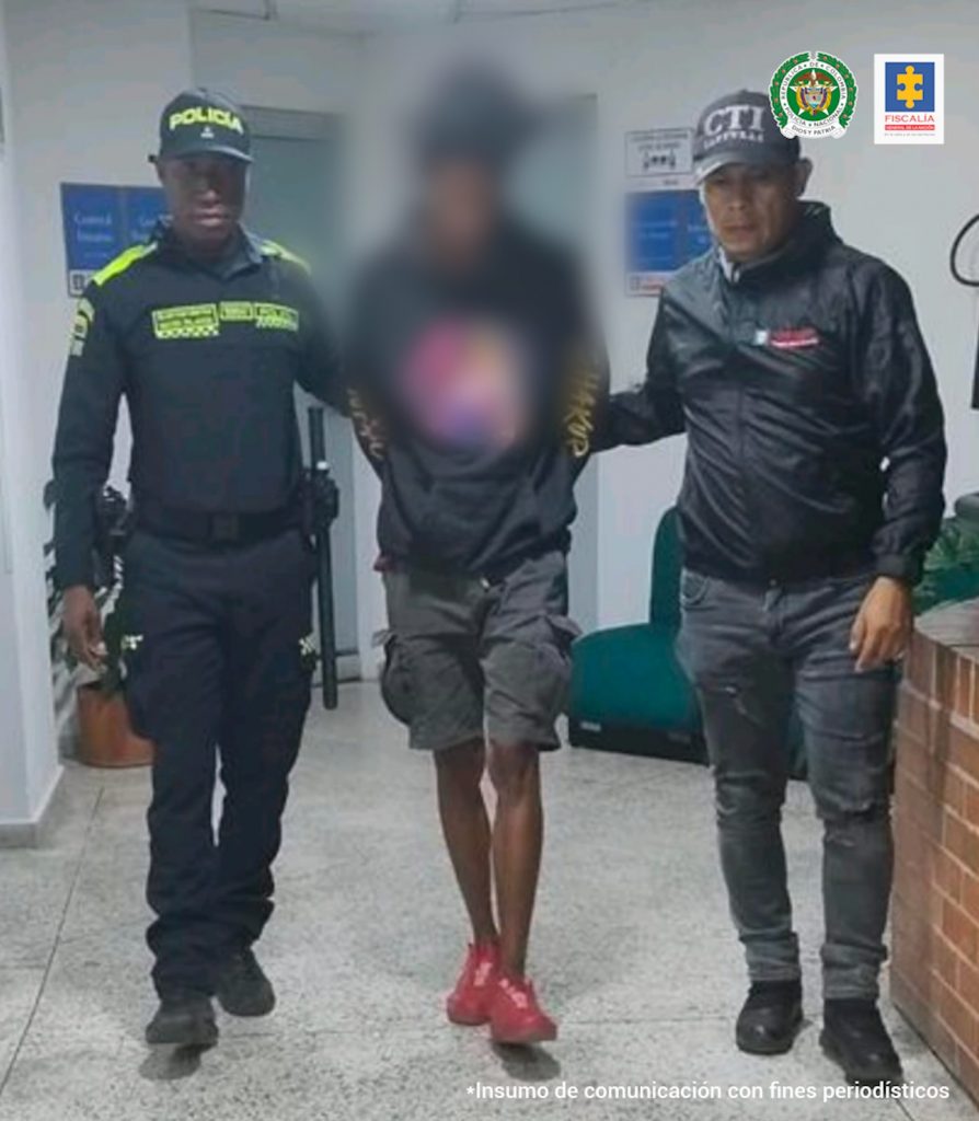 En la fotografía se aprecia al hoy imputado en acción de caminata por un pasillo. Viste buso negro con estampado de colores a la altura del pecho; bermuda tipo cargo gris y zapatillas rojas. Al lado izquierdo de él hay un servidor del CTI de la Fiscalía y al lado derecho un uniformado de la Policía Nacional.