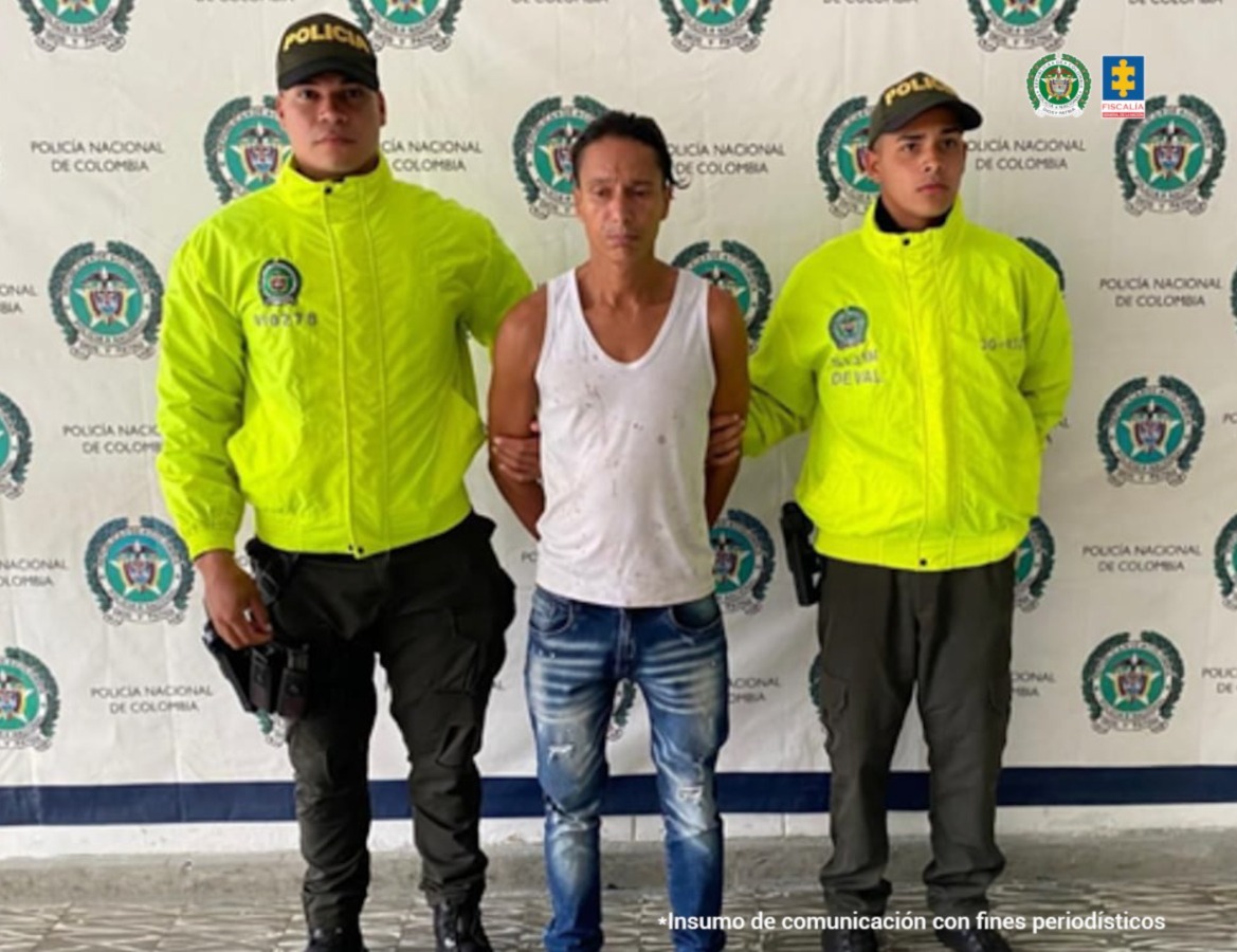 En la fotografía se aprecia de pie y con los brazos atrás a Walter Alonso Picón. Viste con camiseta esqueleto de color blanco, jean azul desgastado y pantuflas grises. A sus extremos hay uniformados de la Policía Nacional. Detrás de ellos hay un pendón institucional de la Policía.