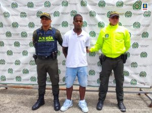 Al centro se observa el capturado y a cada costado un funcionario de Policía Nacional. Al fondo un pendón de Policía Nacional