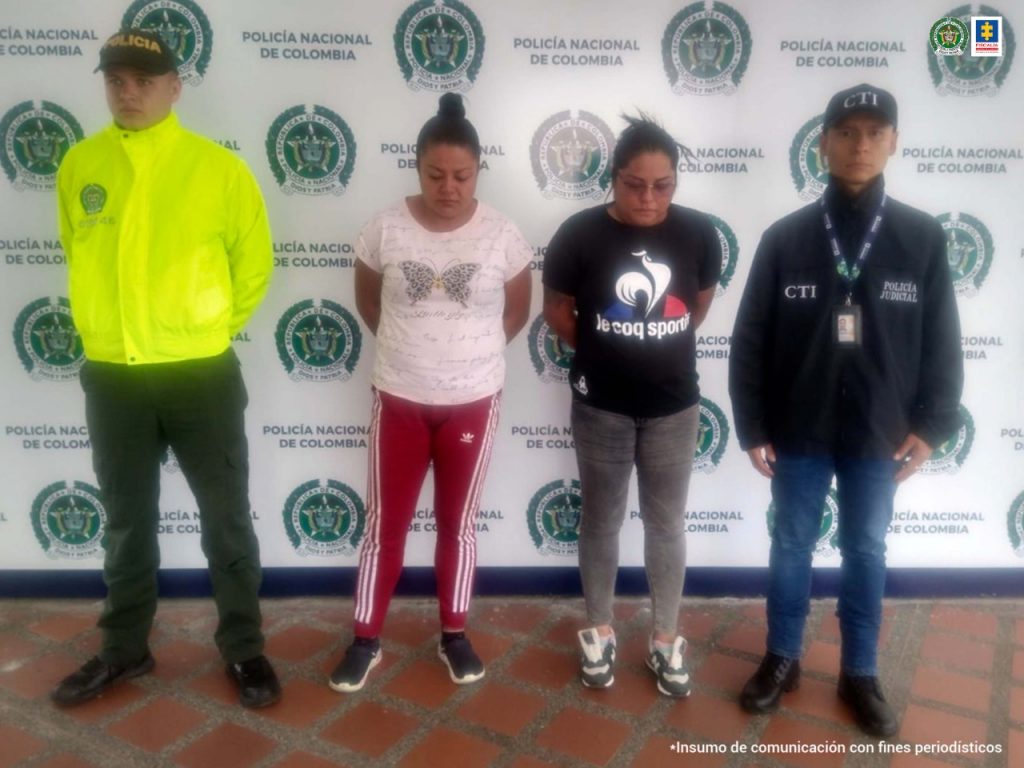 Se observan dos mujeres capturadas. A un costado un funcionario del CTI y al otro uno de Policía Nacional. Al fondo pendón de Policía Nacional