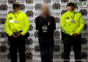 En la foto se ve una persona detenida, custodiada por dos integrantes de la Policía Nacional. Detrás suyo un backing de la institución.