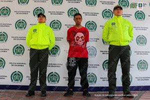 En el centro se observa al capturado y a cada lado un funcionario de Policía Nacional. Al fondo un pendón de Policía Nacional.