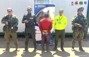El hombre está de espalda a la cámara, está esposado a su espalda y está custodiado por uniformados de la Policía, el Ejército y la Armada Nacional.