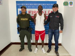 En la foto aparece, de pie y con los brazos atrás, Álvaro Enrique Calderón Izquierdo. Vista camisa negra de manga corta y cuello, pantalón kaki y gorra verde. Al lado derecho de él hay un uniformado de la Policía y al lado izquierdo hay un uniformado del Ejército. Detrás de ellos hay dos pendones institucionales de la Policía y Ejército.