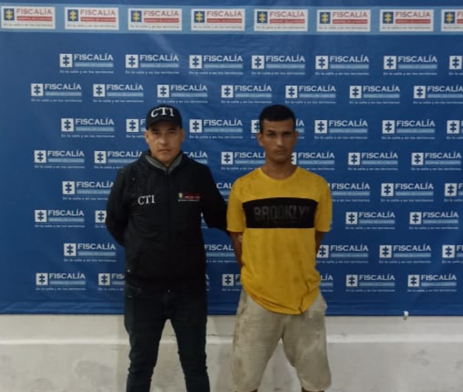 Cristian Camilo Chávez Álvarez esta en la foto acompañado de un funcionario del Cuerpo Técnico de Investigación (CTI). Al fondo un backing con logos de la Fiscalía.