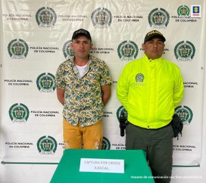 Deimer Jiménez Amaya acompañado de un funcionario de la Policia Nacional y atrás un backing con los logos de la misma institución. Una mesa adelante con mantel verde que dice capturado por orden judicial.
