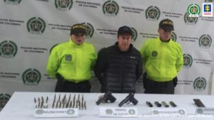 En la fotografía se observa al capturado junto a dos agentes de la Sijin de la Policía Nariño. En la parte posterior se observa una mesa con dos pistolas, 4 proveedores, munición y un teléfono celular