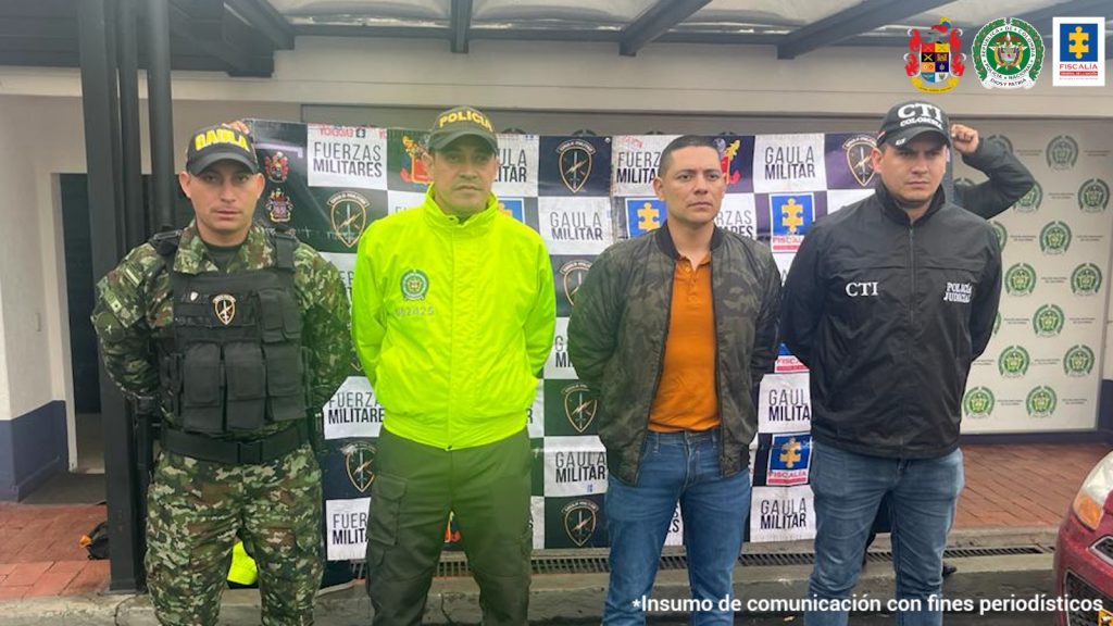 Se aprecia uno de los imputados de pie, con los brazos atrás. Viste chaqueta oscura, camiseta color naranja y jean azul. Al lado izquierdo hay un servidor del CTI de la Fiscalía y al lado derecho uniformados de la Policía y Ejército Nacional. Detrás de ellos hay un pendón interinstitucional con logos del Ejército, Policía y Fiscalía.