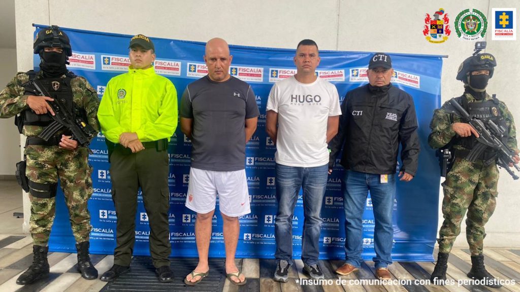 Se aprecian dos de los hoy imputados, de pie y con los brazos atrás. Uno de ellos vista camiseta oscura, pantaloneta blanca y pantuflas. La otra persona viste camiseta de manga corta blanca, jean azul zapatos oscuros. A la izquierda de ellos hay un servidor del CTI de la Fiscalía y un uniformado del Ejército Nacional. A la derechos de ellos hay uniformados de la Policía y Ejército Nacional. Detrás de ellos hay un pendón institucional de la Fiscalía.