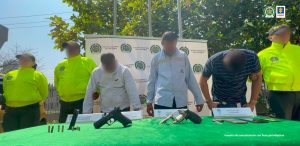 En la imagen aparecen tres hombres de camisa blanca, camisa azul clara y camiseta azul oscura de rallas blancas, custodiados por tres agentes de la Policía Nacional. Delante de ellos se observa una mesa con armas y cartuchos incautados.