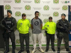 En la foto aparece, de pie Johan Alberto Brand López. Viste camiseta gris de manga corta con logo circular en negro; pantalón color beage y tenis claros. A lado y lado de él hay uniformados de la Policía Nacional. Detrás de ellos hay un pendón institucional de la Policía.