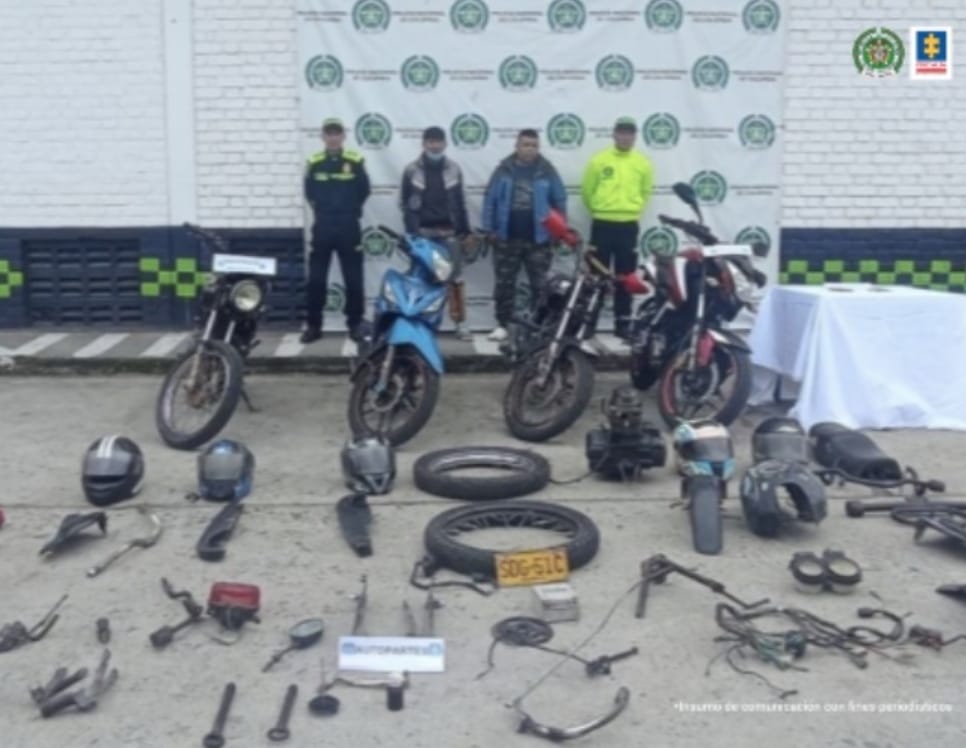 En la fotografía se observa a los dos capturados junto a dos agentes de la Policía Nacional. En la parte posterior se observan cuatro motocicletas, partes de motos, cascos, llantas, entre otros elementos. 