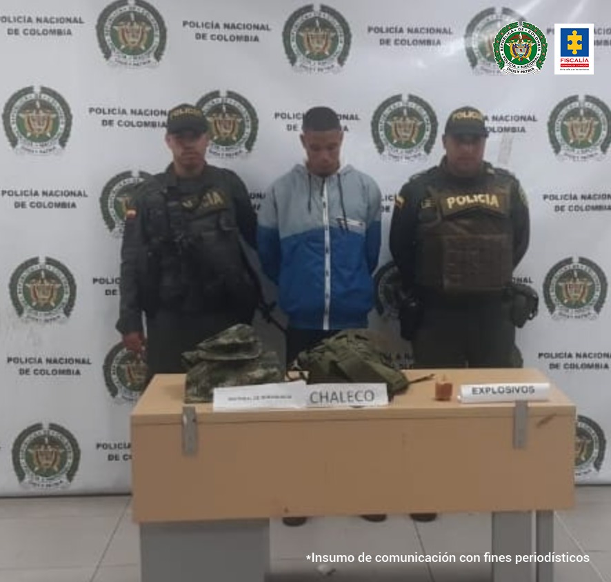 En la fotografía se observa al capturado junto a dos agentes de la Policía Nacional. En la parte posterior se observa una mesa con una chaqueta pixelada del Ejército, un arnés y un explosivo