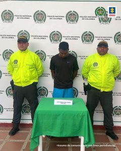 En la imagen se observa a un hombre vestido de camiseta negra con pantalón azul claro, custodiado por dos agentes de la Policía Nacional delante de un pendón de esa institución.
