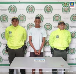 En la fotografía aparece un hombre delgado de tez morena, con camiseta gris y bermuda café, custodiado por dos uniformados de la Policía Nacional delante de un pendón de esa institución.