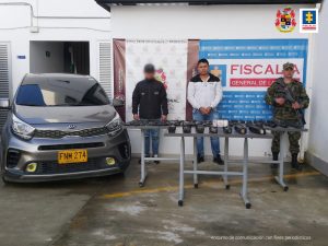 En la imagen se aprecia a Yeison David Arias López quien está escolado a lado y lado de su cuerpo por uniformados del CTI y del Ejército. En frente sobre una mesa reposan el dinero que fue encontrado escondido en un vehículo particular que conducía. A mano derecho del procesado se observa el vehíuculo incautado. 