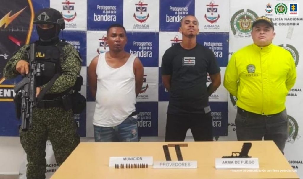 En la fotografía se observa a los dos capturados junto a un agente de la Policía Nacional y a un soldado del Gaula Militar. En la parte posterior se observa una mesa en la que está colocada un arma de fuego y munición