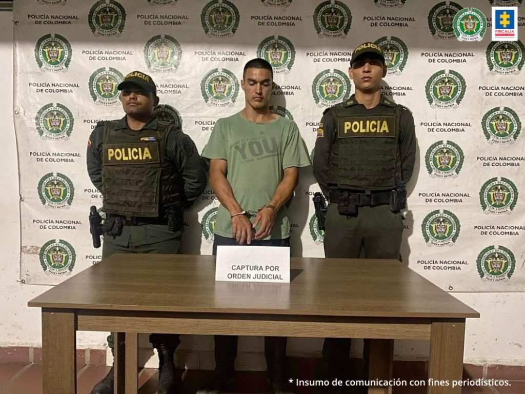 En la foto aparece Sebastián Vera Montealegre acompañado de dos funcionarios de la Policía Nacional a los lados, una mesa café adelante y atrás un backing con los logos de la Policía.