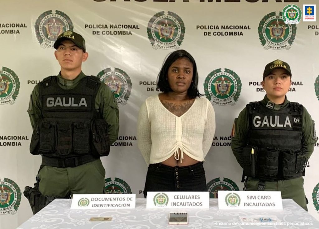 En la fotografía aparece la capturada junto a dos agentes del Gaula de la Policía Nacional. En la parte posterior, se observa una mesa en donde está una cédula, un teléfono celular y 6 simcard