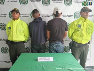 Jhon Anderson Pestana Alvarán, alias Galindo; y Jaider Valencia aparecen en la foto de espaldas custodiados por funcionarios de la Policía Nacional a los lados. Al fondo un pendón con logos de la Policía y adelante una mesa con mantel verde.