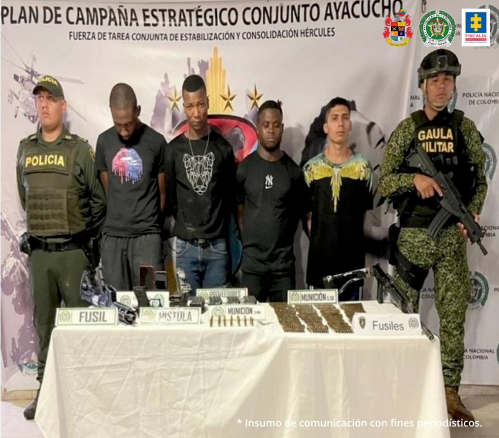 En la fotografía se observan a los cuatro capturados junto a un agente de la Sijn de la Policía Nacional y un soldado del Gaula Militar. En la parte posterior se observa una mesa con armamento, munición y dos teléfonos celulares