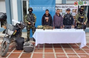 En la fotografía se observa a los dos capturados junto a un servidora del CTI y dos solados del Gaula Militar. En la parte superior se observa una mesa con dos armas, munición, uniformes del Ejército Nacional, un chaleco con arnés, una motocicleta y dos maletines