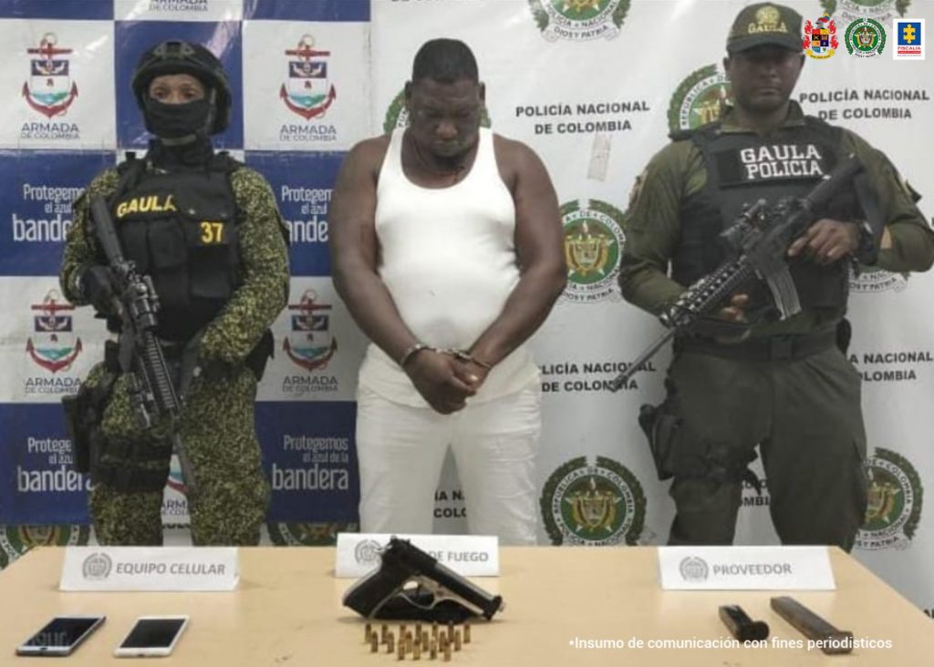 En la fotografía se observa al capturado junto a un agente del Gaula de la Policía y un soldado del Gaula Militar. En la parte posterior se observa una mesa con un arma de fuego, munición, dos proveedores y dos teléfonos celulares