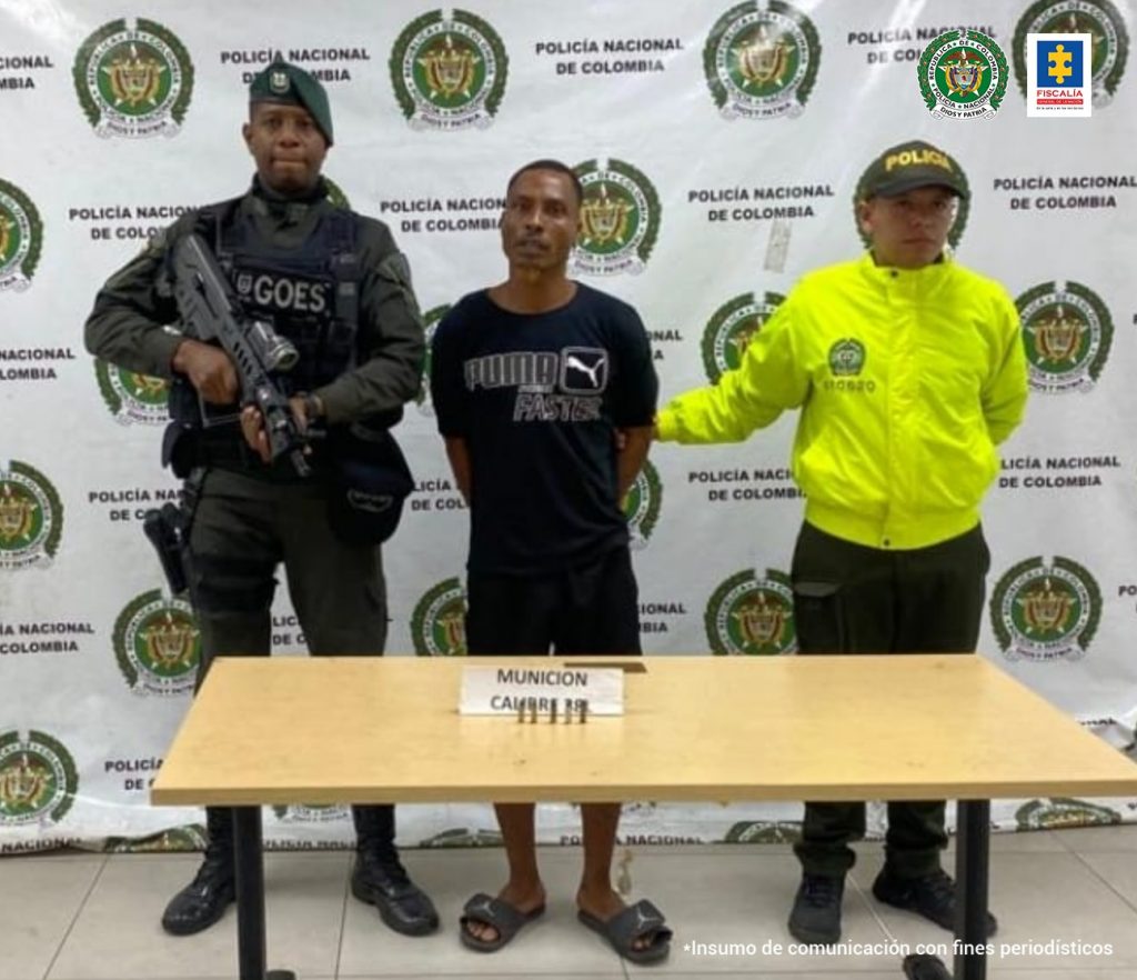 En la fotografía se observa al capturado junto a un agente del Gaula de la Policía y un soldado del Gaula Militar. En la parte posterior se observa una mesa con un arma de fuego, munición, dos proveedores y dos teléfonos celulares
