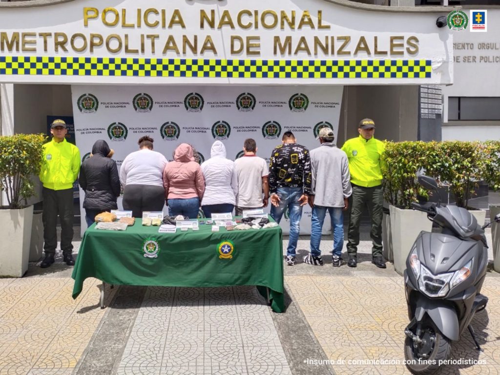 En la foto aparecen los 7 detenidos de espaldas acompañados por funcionarios de la Policía a los lados. Adelante una mesa con mantel verde con los elementos incautados.