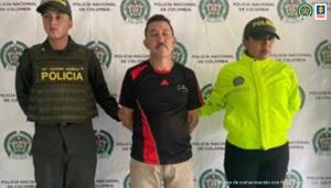 En la fotografía aparece el capturado junto a un servidor del CTI y a un agente de la Policía Nacional. En la parte superior está un banner de la Fiscalía General de la Nación y un banner de la Policía Nacional
