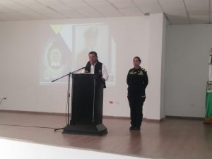  En la imagen está un hombre hablando detrás de un atril, vestido con una chaqueta negra que dice Fiscalía General de la Nación. A su lado aparece una mujer con uniforme de la Policía Nacional. 