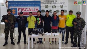 En la imagen se aprecia a los capturados junto a un investigador del CTI y uniformados del Ejército y Policía Nacional. En la parte posterior se observan los banners que identifican a la Fiscalía General de la Nación y a la Policía Nacional.