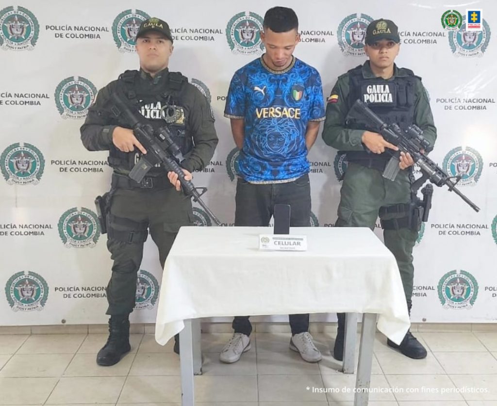 En la fotografía aparece Nick Kevin Tenorio Prado junto a dos agentes del Gaula de la Policía Nacional. En la parte posterior está una mesa con un teléfono celular
