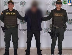 En la fotografía está el capturado junto a dos agentes de la Policía Nacional. En la parte superior se observa un banner de la Fiscalía General