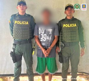 El capturado está custodiado por dos policías y con sus manos al frente y esposadas.