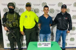 En la fotografía aparece el capturado junto a un servidor del CTI y dos agentes de la Policía Nacional. En la parte superior está un banner de la Policía Nacional