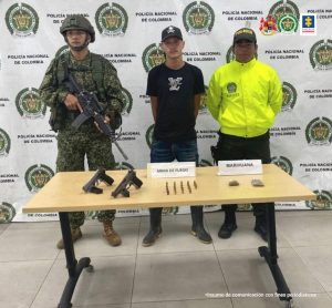 En la fotografía aparece el capturado junto a un infante de la Armada Nacional y un agente de la Policía Nacional. En la parte posterior está una mesa con dos armas de fuego, munición y estupefacientes