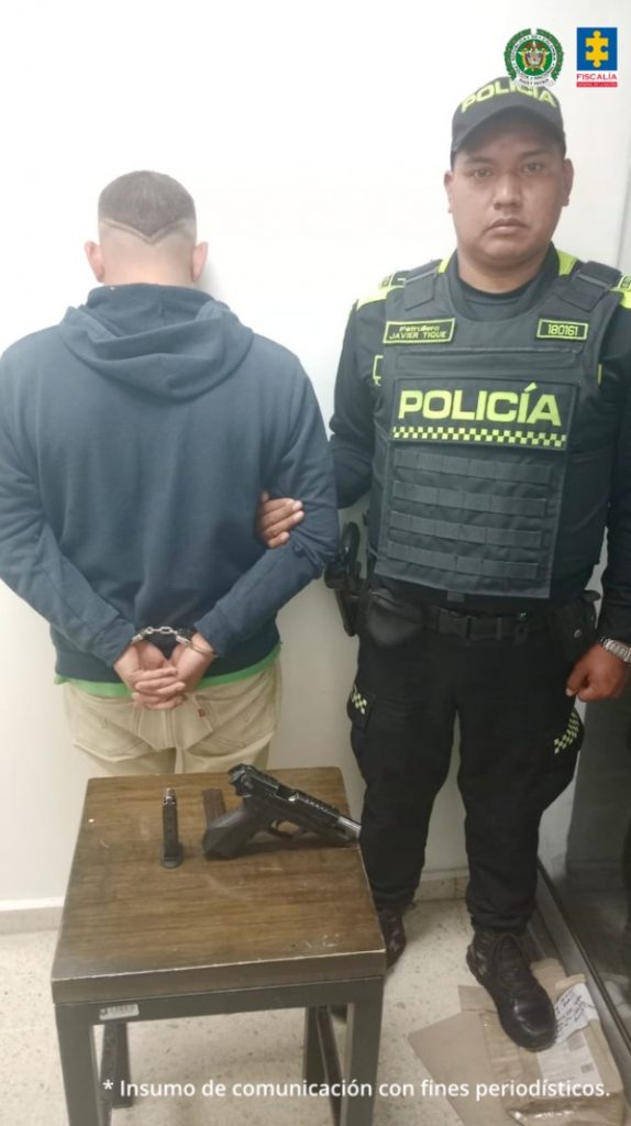En la imagen un hombre de espaldas acompañado de un policía al frente de ellos una mesa con un arma encima de la misma.