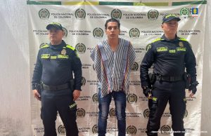 El imputado se encuentra en custodia por dos uniformados de la Policía Nacional. Detrás se observa un backing de la Policía.