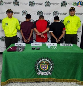 en la imagen dos policías y en el medio tres capturados al frente una mesa con un mantel verde donde se observa prendas decomisadas, celulares, cocaína y munición
