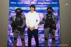 Se ve al capturado, Felipe Isaza Cadavid con dos agentes del grupo táctico del CTI de la Fiscalía que lo custodian a lado y lado. Al fondo un backing de la Fiscalía General.