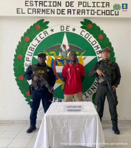 El procesado aparece con camiseta roja de pie acompañado de dos uniformados. De fondo se observa un pendón de la Policía Nacional