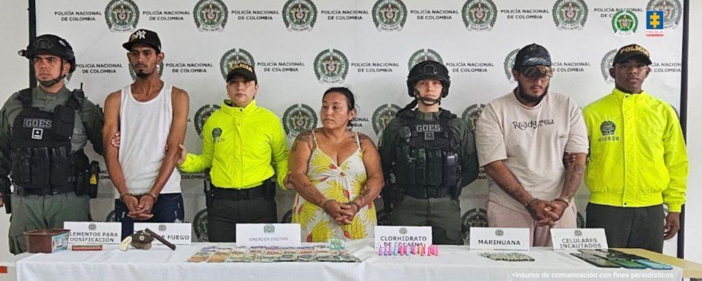 Los capturados aparecen esposados y custodiados por uniformados de la Policía, en una mesa frente a ellos, los elementos incautados durante el allanamiento tales como un arma de fuego, estupefacientes y dinero en efectivo.