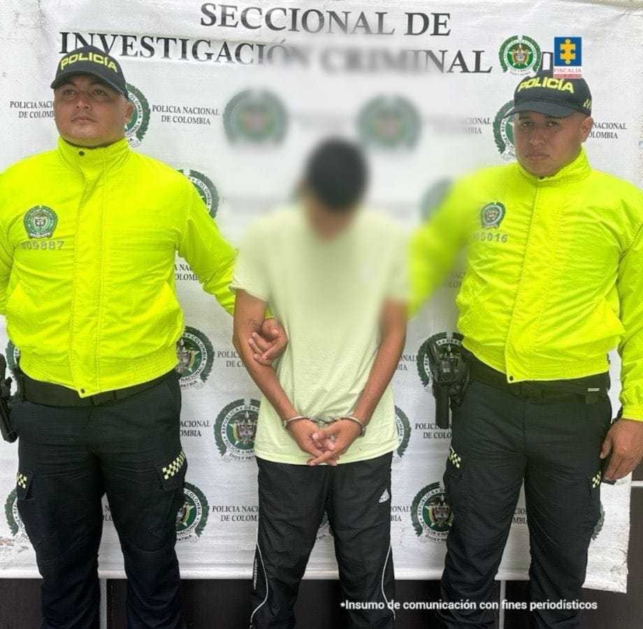  Se visualiza al capturado junto a dos uniformados de la Policía Nacional. Detrás de ellos el banner que identifica a la Policía Nacional.