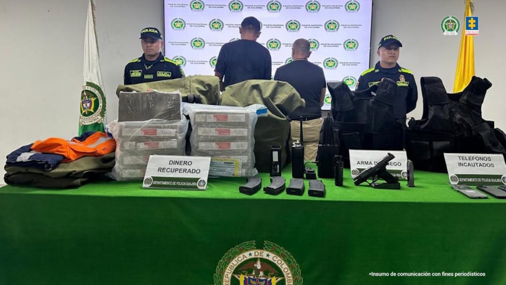 En la imagen se ve una mesa cubierta con un mantel verde con el escudo de la República de Colombia. Encima de la mesa hay diferentes objetos incautados por la Policía Nacional y la Fiscalía, organizados por categorías. A la izquierda, hay ropa doblada, posiblemente uniformes o prendas usadas para cometer delitos. En el centro, se encuentran varios paquetes de dinero etiquetados como “DINERO RECUPERADO”, apilados y envueltos en plástico. También hay radios de comunicación, cargadores de armas, y un arma de fuego etiquetada como “ARMA DE FUEGO”.A la derecha, hay chalecos antibalas negros y varios teléfonos celulares bajo el cartel “TELÉFONOS INCAUTADOS”. Detrás de la mesa hay cinco personas: dos policías uniformados en los extremos y tres personas de civil, de espaldas. Al fondo, se ve una pantalla con los logos de la Policía Nacional y de la Fiscalía. A los lados hay dos banderas: la bandera de Colombia y otra institucional. La escena corresponde a una presentación oficial de resultados operativos, y abajo a la derecha se lee: “Insumo de comunicación con fines periodísticos”.
