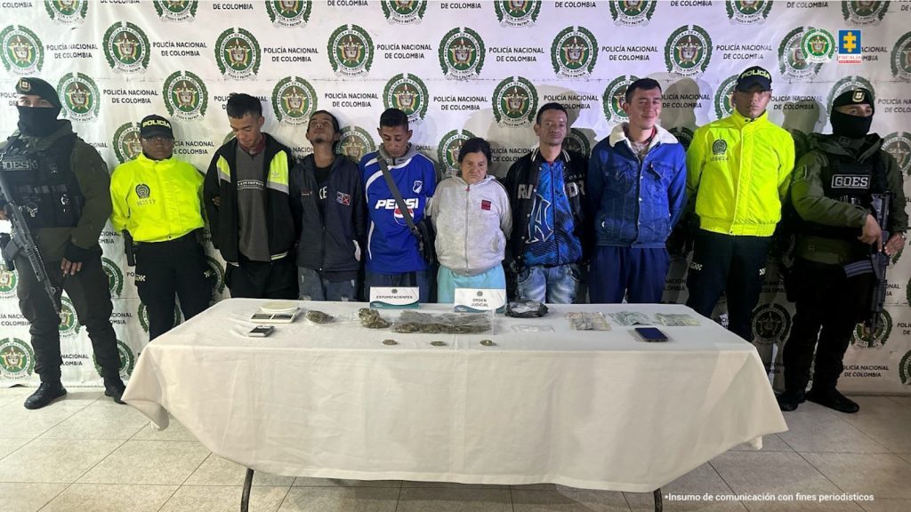 en la imagen se pueden ver a los 6 capturados en medios de 2 policias y 2 agentes del GOES, atras de ellos se ve un baking de la policia y en frente de ellos hay una mesa con los estupefacientes incautados