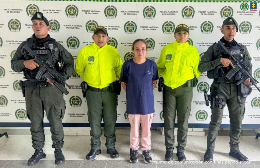 En la imagen se ve a una mujer en el centro, esposada y custodiada por cinco agentes de la Policía Nacional de Colombia. Ella viste una camiseta azul oscura, pantalón rosado claro y zapatillas negras. Tiene el cabello recogido y una expresión seria. A su lado, dos policías visten chaquetas amarillas reflectivas con el logotipo de la Policía, mientras que los otros tres, ubicados en los extremos izquierdo y derecho, llevan uniformes oscuros y están armados, del grupo GOES (Grupo de Operaciones Especiales). Todos posan frente a un fondo institucional con los logotipos repetidos de la Policía Nacional de Colombia. En la esquina superior derecha aparece también el escudo de la Fiscalía General de la Nación.